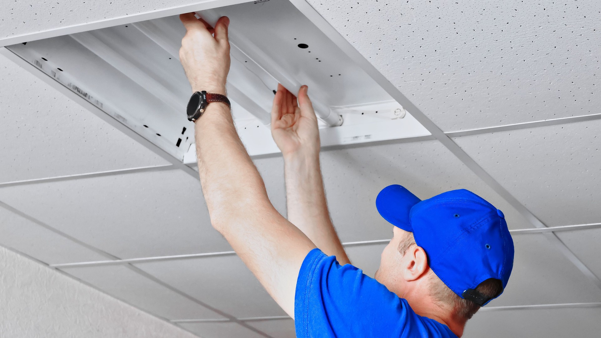 Electrician fixing ceiling fluorescent light panel in office building using tools.